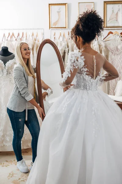 Tailor stands next to the mirror in a bridal salon and helps the bride choose her wedding dress.