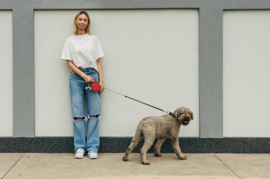 Önde genç sarışın bir kadının tasmalı köpeğiyle kaldırımda duruşu.