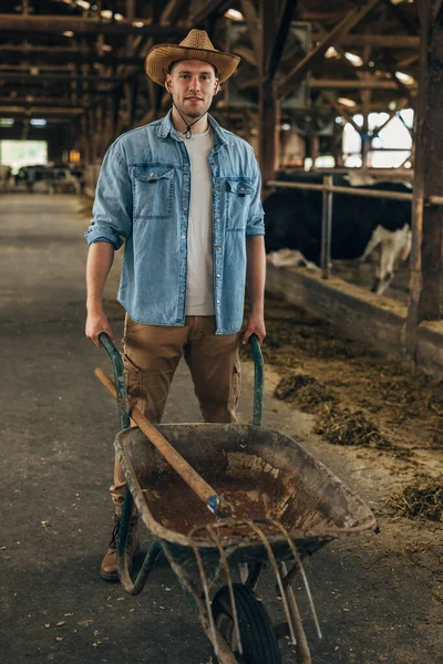 Çiftçi ahırda duruyor ve içinde saman çatalı olan bir arabayı itiyor..