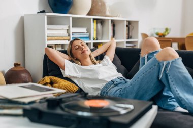 Beyaz kadın evde gramofondan müzik eşliğinde dinleniyor.