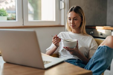 Genç bir kadın mutfakta yulaf ezmesi yiyor ve dizüstü bilgisayarına bakıyor..