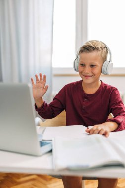 Genç bir erkek öğrenci çevrimiçi sınıfta kameraya el sallıyor..