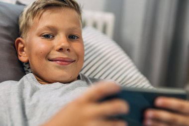 Tatlı küçük çocuk yatakta uzanıyor ve cep telefonunu kullanıyor..