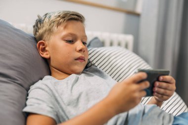 Beyaz bir çocuk yatakta uzanıyor ve akıllı telefonunu kullanıyor.