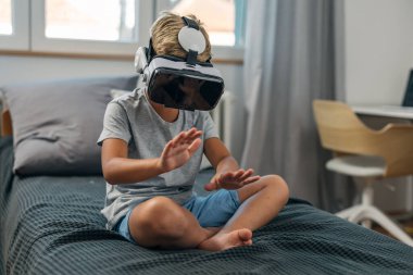 Genç çocuk yatakta oturuyor ve sanal gerçeklik gözlükleriyle oyun oynuyor..