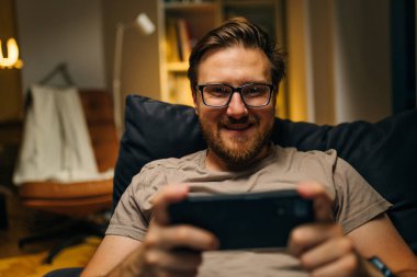 Mutlu beyaz adam evde telefonunu kullanıyor ve kameraya bakıyor..