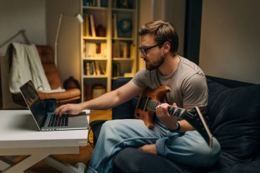 Beyaz bir adamın evde oturup gitar çalışmasının yan görüntüsü..