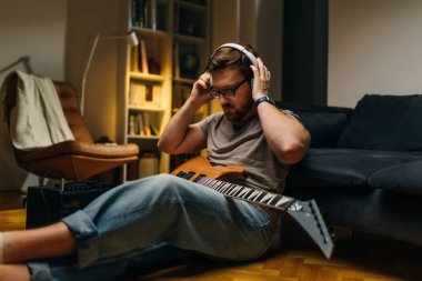 Evde yerde elektro gitarla oturan ve kulaklık takan bir müzisyen..