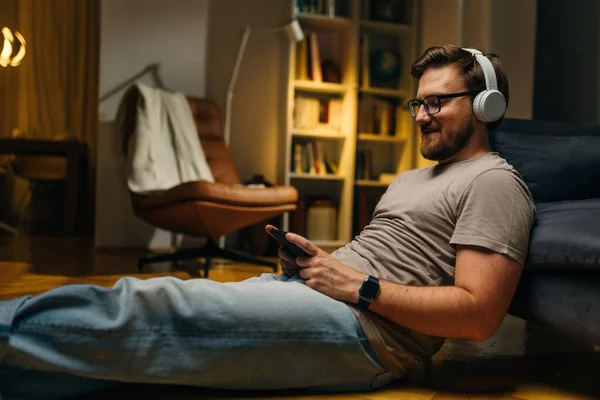 Side view of a relaxed man sitting on he floor in the living room and using mobile phone to play music.
