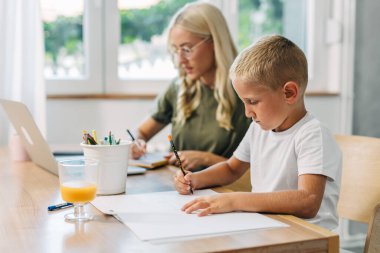 Annesi yanında dizüstü bilgisayarla çalışırken küçük tatlı bir çocuk resim çiziyor..