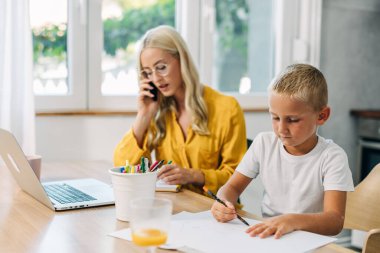 Sevimli beyaz çocuk evde çalışan annesinin yanında ev ödevi yapıyor..