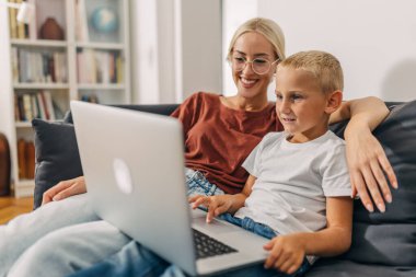 Beyaz çocuk evinde annesinin yanında dizüstü bilgisayar kullanıyor..