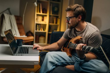 Elektro gitarlı beyaz bir adamın dizüstü bilgisayardaki notlara bakması.