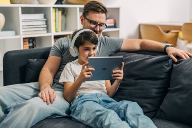 Genç bir çocuk babasıyla kanepede oturup dijital tablet kullanarak video oyunu oynuyor..