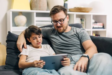 Kanepede oturup dijital tablet kullanan beyaz bir baba ve oğul ön görüntüsü..