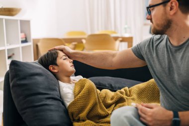 Bir çocuk battaniyeyle kaplı bir kanepede hasta yatıyor, babası ateşini kontrol ediyor..