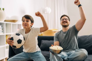 Genç bir çocuk ve babası favori takımları için tezahürat yapıyorlar ve birlikte futbol maçı izliyorlar..