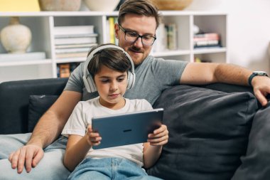 Genç çocuk tablet ve kulaklık kullanıyor ve babası da onunla birlikte oturup ekrana bakıyor..