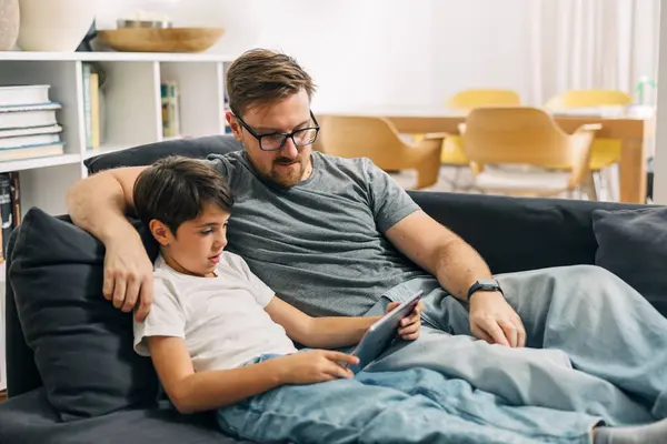 Father and son sitting together on a couch and watching motion picture on tablet.