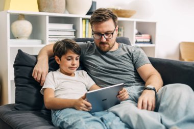 Baba ve oğul bir kanepede dinleniyorlar ve dijital tablet kullanıyorlar..