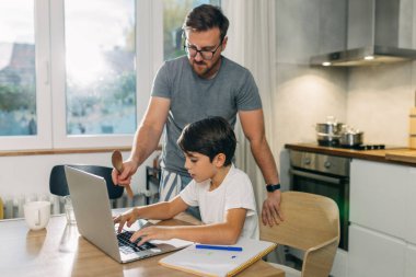 Baba yemek pişirirken oğluna laptopu kullanmasında yardım ediyor..