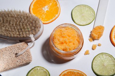 An orange scrub in a glass jar on a white table with pieces of citrus fruits. Nearby is a brush made of natural bristles for body peeling, a pumice stone for feet. clipart