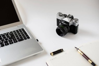 Gri bir dizüstü bilgisayarın yanında beyaz bir masanın üzerinde duran eski bir eski model fotoğraf makinesinin lensi ve kayıt için bir not defteri..