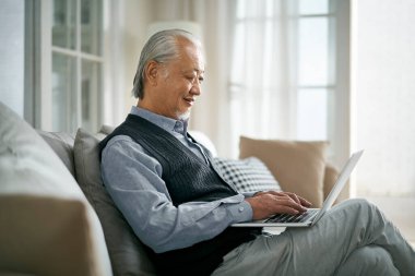 Evde dizüstü bilgisayar kullanarak koltukta oturan yaşlı Asyalı adamın yan görüntüsü.