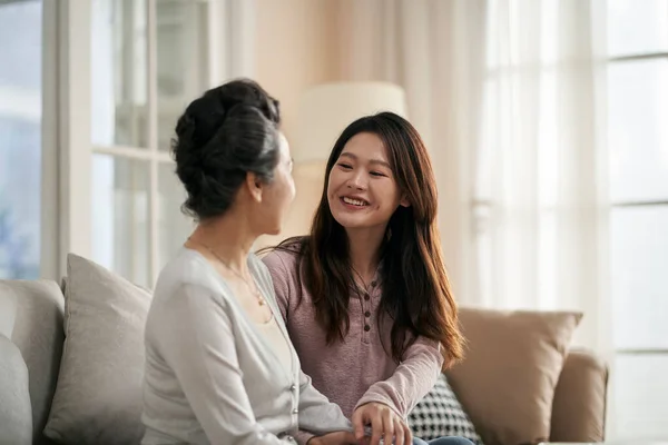 亚洲的成年女儿坐在家里的沙发上 与年长的母亲谈天说地 笑容满面 — 图库照片