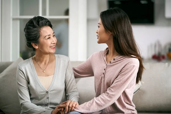 亚洲的成年女儿坐在家里的沙发上 与年长的母亲谈天说地 笑容满面 — 图库照片