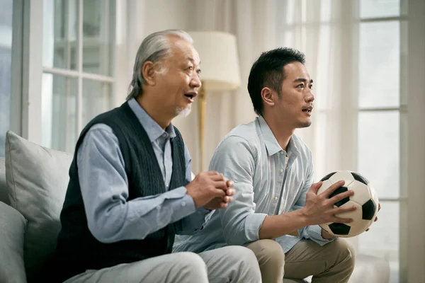 年长的亚裔父亲和成年儿子一起坐在家里的沙发上看电视足球赛 — 图库照片