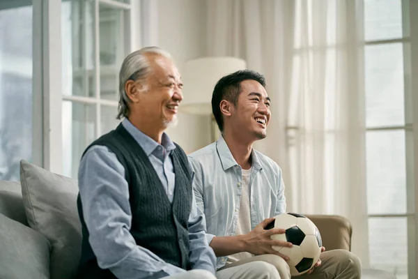 年长的亚裔父亲和成年儿子一起坐在家里的沙发上看电视足球赛 — 图库照片