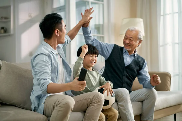 asian son father grandfather sitting on couch at home celebrating goal and victory while watching live broadcasting of football match on TV together