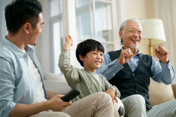 亚洲儿子的父亲爷爷坐在家里的沙发上 一边庆祝胜利和进球 一边观看电视直播足球比赛 — 图库照片