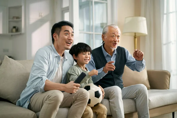 asian son father grandfather sitting on couch at home watching live broadcasting of football match on TV together