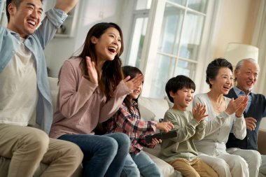 multi generational asian family sitting on couch in living room at home watching sports game on tv