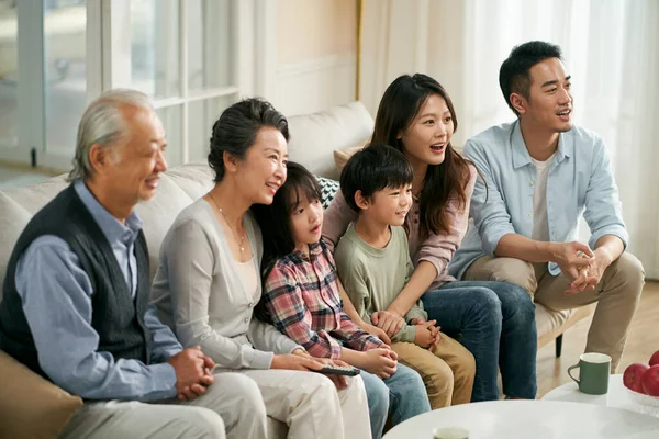 three generation asian family sitting on couch at home watching tv together happy and smiling