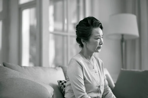 side view of sad senior asian woman sitting alone on couch in living room at home, black and white