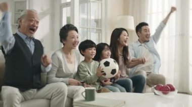 three generation asian family sitting on couch at home watching live broadcasting of soccer game together celebrating a goal