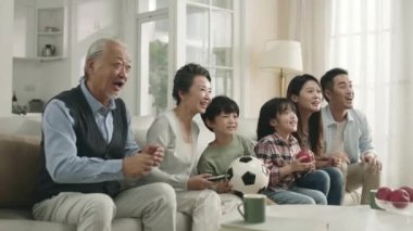 Üç kuşak Asyalı aile evlerinde oturmuş futbol maçının canlı yayınını izliyorlar ve bir golü kutluyorlar.