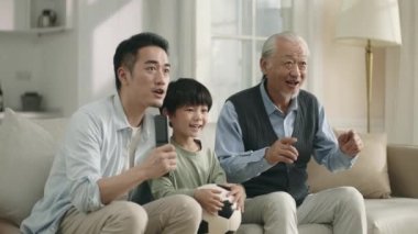 asian son father and grandfather watching live broadcasting of football match on TV together at home celebrating a goal