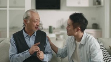 asian adult son sitting on couch at home conversing with senior father happy and smiling