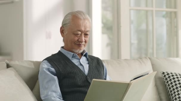 Starszy Azji Człowiek Siedzi Kanapie Domu Czytając Książkę — Wideo stockowe