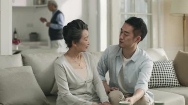 young asian adult son sitting on couch chatting with senior mother during visit of parents