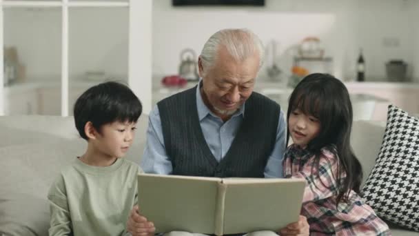 Asian Grandpa Two Grandchildren Sitting Couch Home Reading Book Together — Stock video