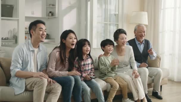 家庭でテレビでスポーツ大会を見ながらソファに座っている3世代のアジア系家庭 — ストック動画