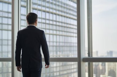 Asyalı iş adamının dikiz aynasından şehir manzarasına bakarak pencereye doğru yürümesi