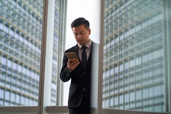 asian businessman looking at mobile phone in office with modern building in background and in glass reflection