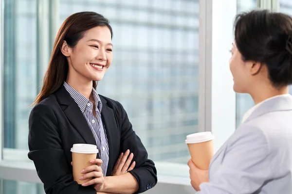 現代のオフィスで窓のそばでおしゃべりをしている若いアジア系ビジネス女性2人 — ストック写真