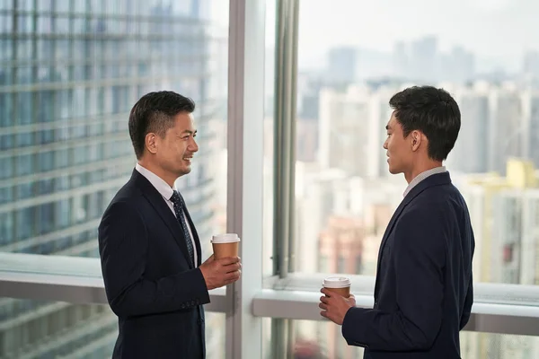 現代オフィスの窓のそばに立つ2人のアジア人ビジネスマンがチャットで話し合い — ストック写真